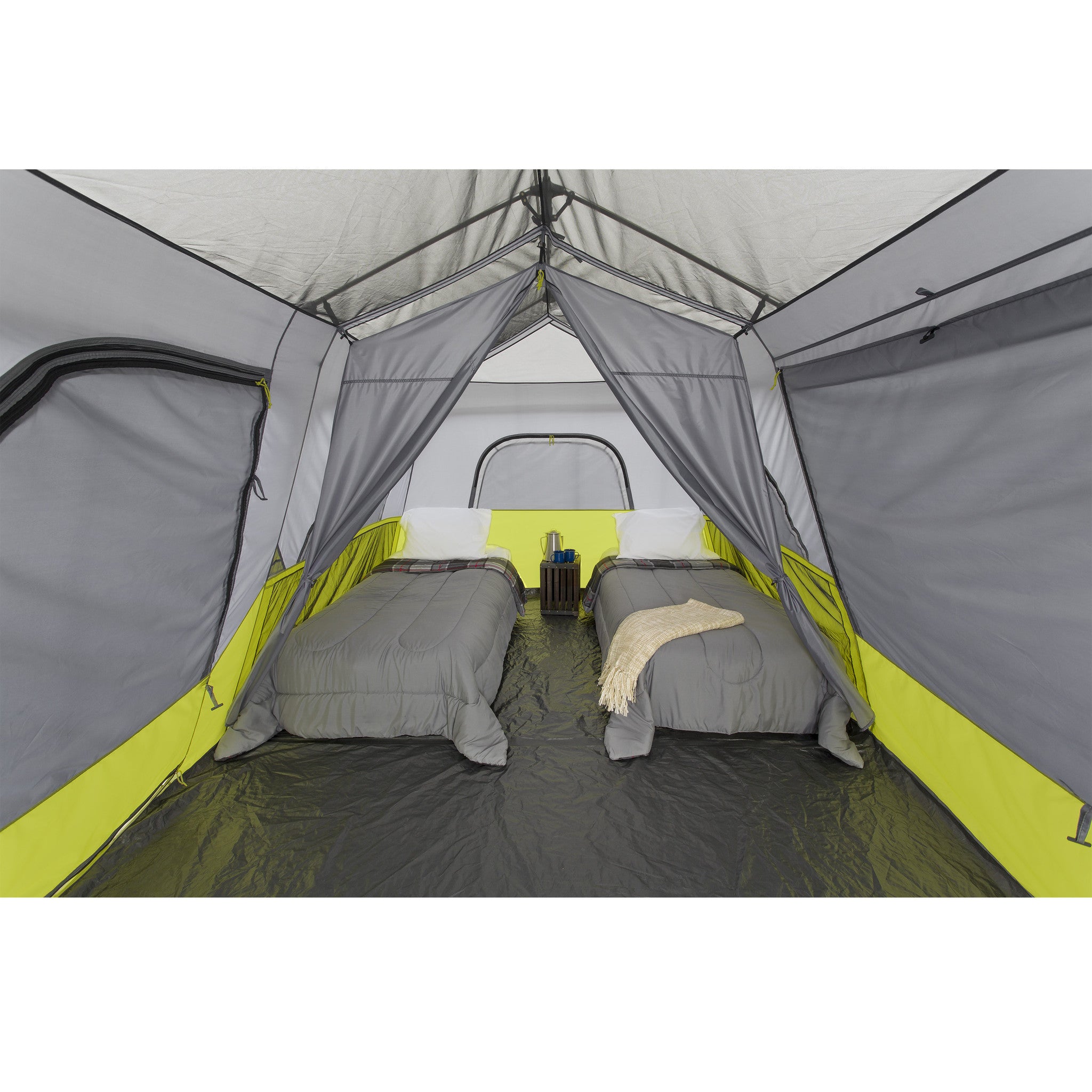 The interior of a spacious CORE 9 Person Instant Cabin Tent featuring two single air mattresses, each with pillows and bedcovers. A small wooden table with a lantern rests between the mattresses, and the tent has large windows and a door with mesh panels for ventilation.