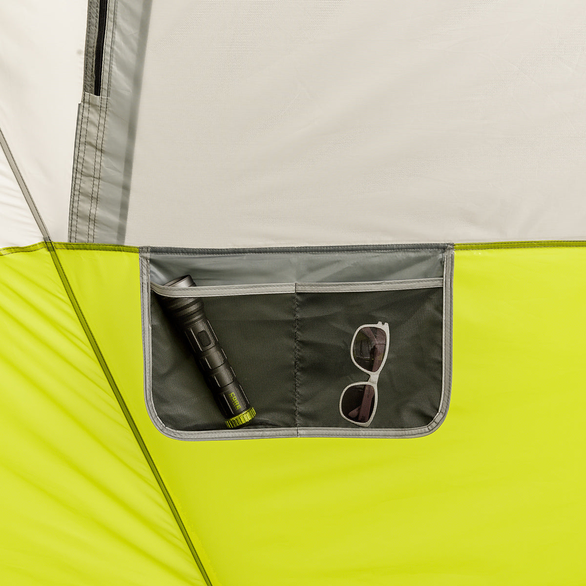 A close-up of a green and gray tent's interior pocket. The pocket holds a black flashlight and a pair of white-framed sunglasses. The tent's fabric is light gray with a green lower section, and the pocket is neatly stitched onto the wall of the CORE 12 Person Instant Cabin Tent.