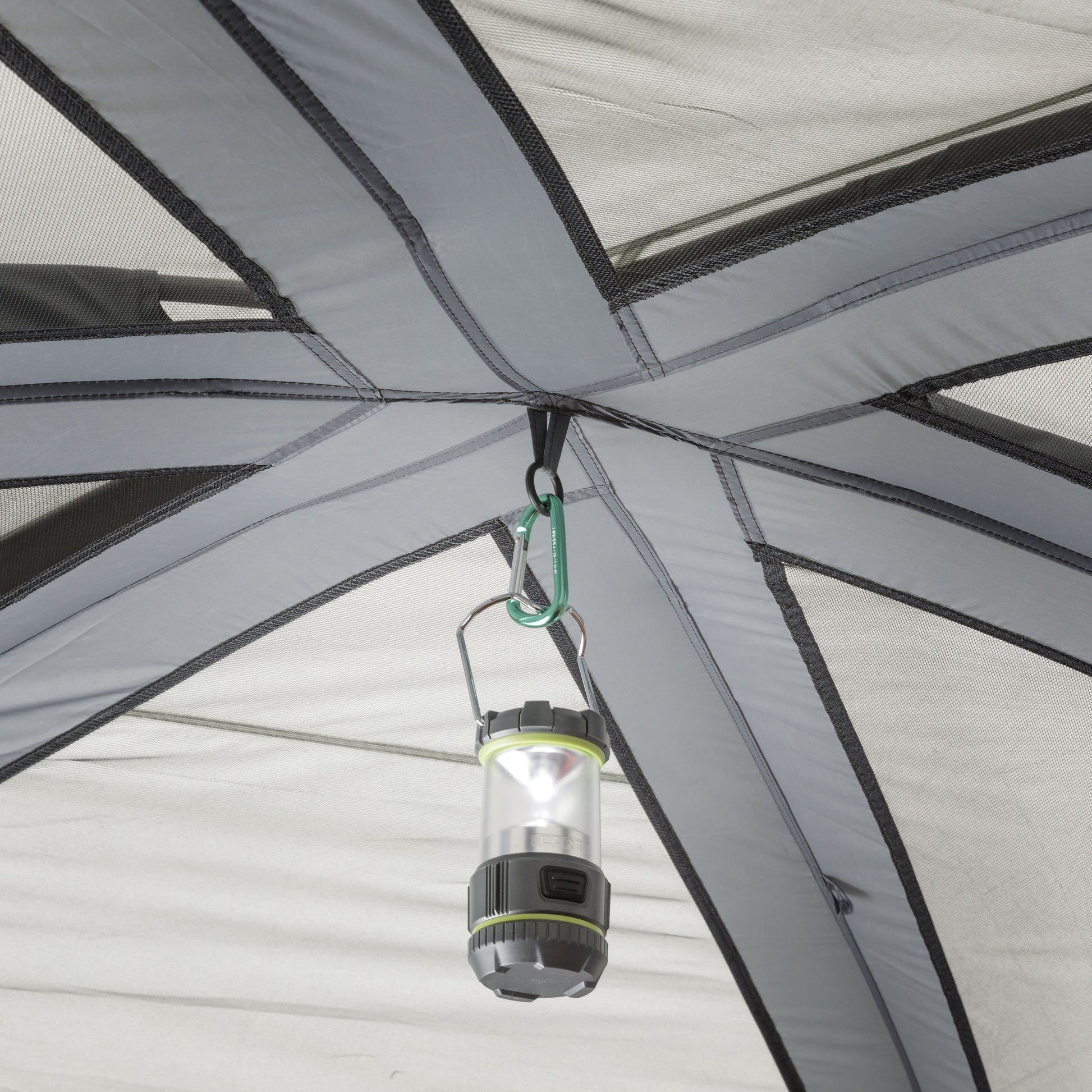A close-up of a grey tent interior shows a small portable lantern hanging from the ceiling. The lantern is attached to the tent's top support structure with a green carabiner. The fabric and poles of the CORE 10 Person Straight Wall Cabin Tent are visible, providing support for the lantern.