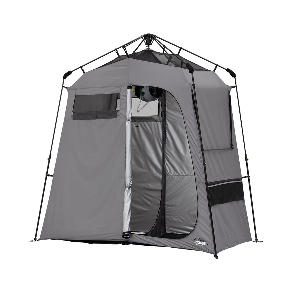 A gray Two Room Instant Shower Tent is set up with its door open. The tent features a sturdy frame, multiple zippered windows for ventilation, and mesh storage pockets on the side. The tent is branded with the name CORE on the bottom right corner.