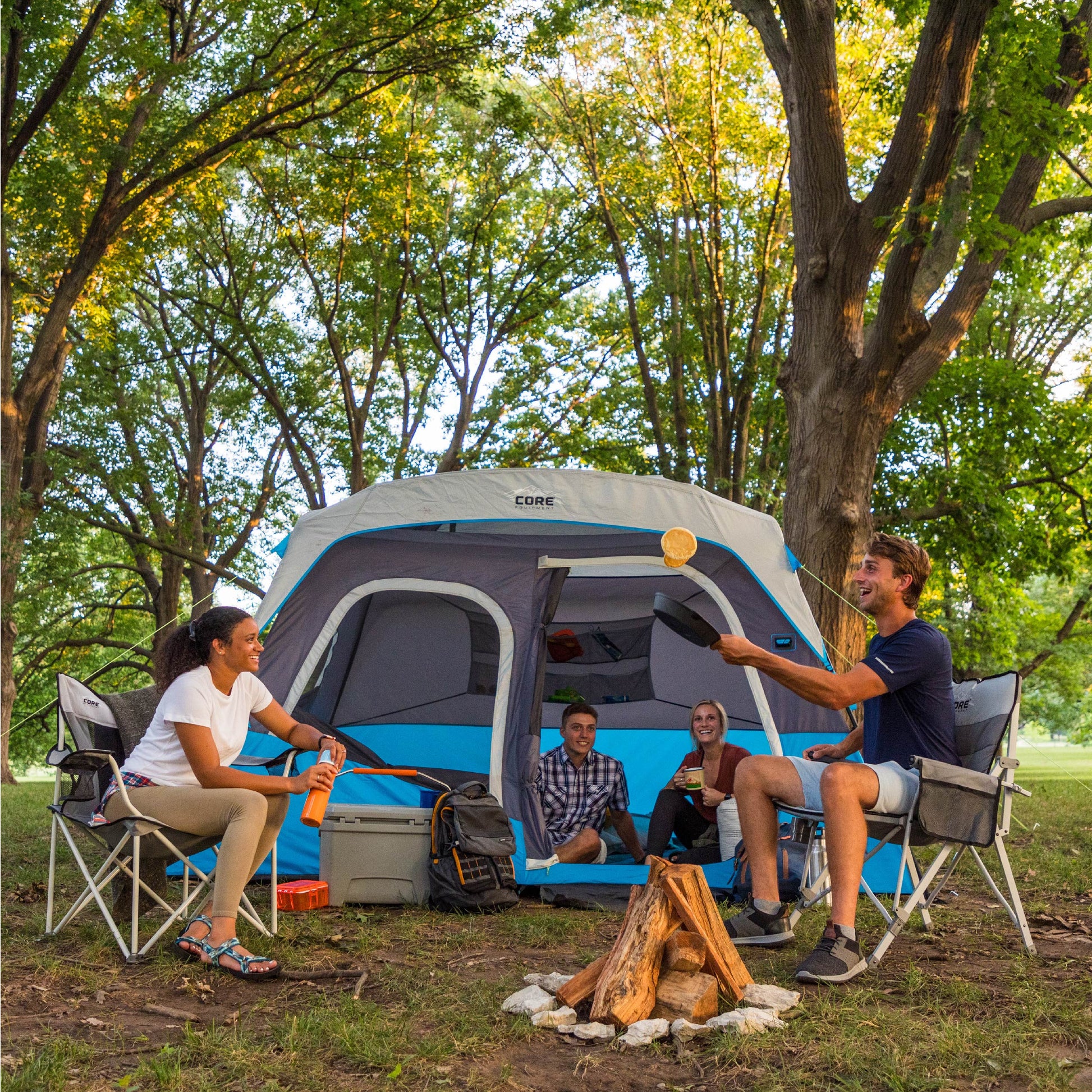 Four people sit around a campfire outside a blue and grey 6 Person Lighted Instant Cabin Tent by CORE in a wooded area. One person is holding a marshmallow on a stick, preparing to toast it. The group appears to be enjoying the outdoor setting, with tall trees and green foliage in the background.