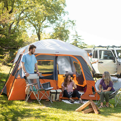 Three people are camping in a grassy area with trees in the background. They are seated beside a CORE 6 Person Straight Wall Cabin Tent, with camping gear like coolers and a kettle around them. One person is standing next to a cooler, while the other two are sitting in camping chairs. A parked SUV with roof racks is visible behind the tent.
