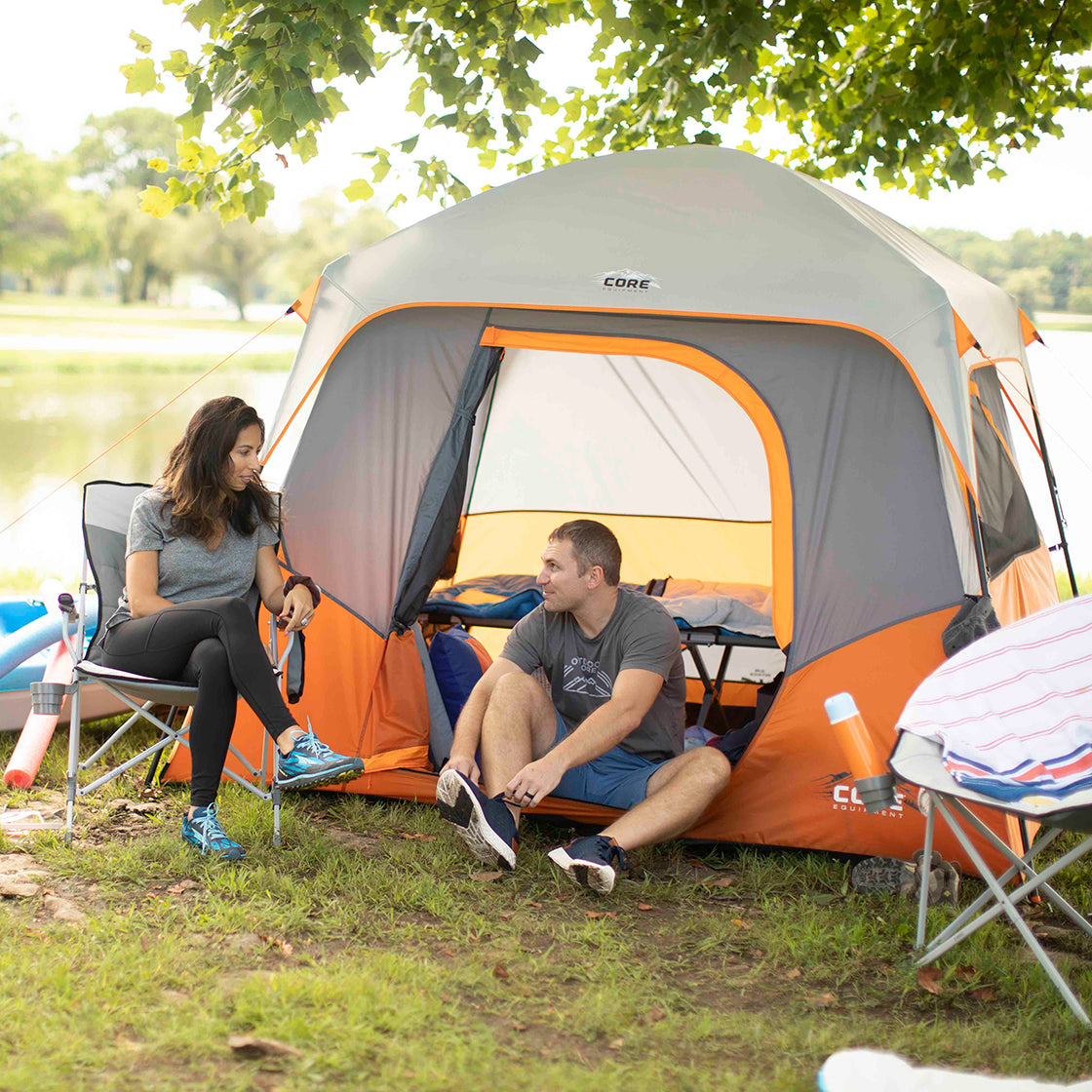 Two people sitting on camping chairs outside a CORE 4 Person Straight Wall Cabin Tent. They are near trees with a lake in the background. One person is tying their shoes while the other is holding a water bottle. Camping gear is scattered around them.