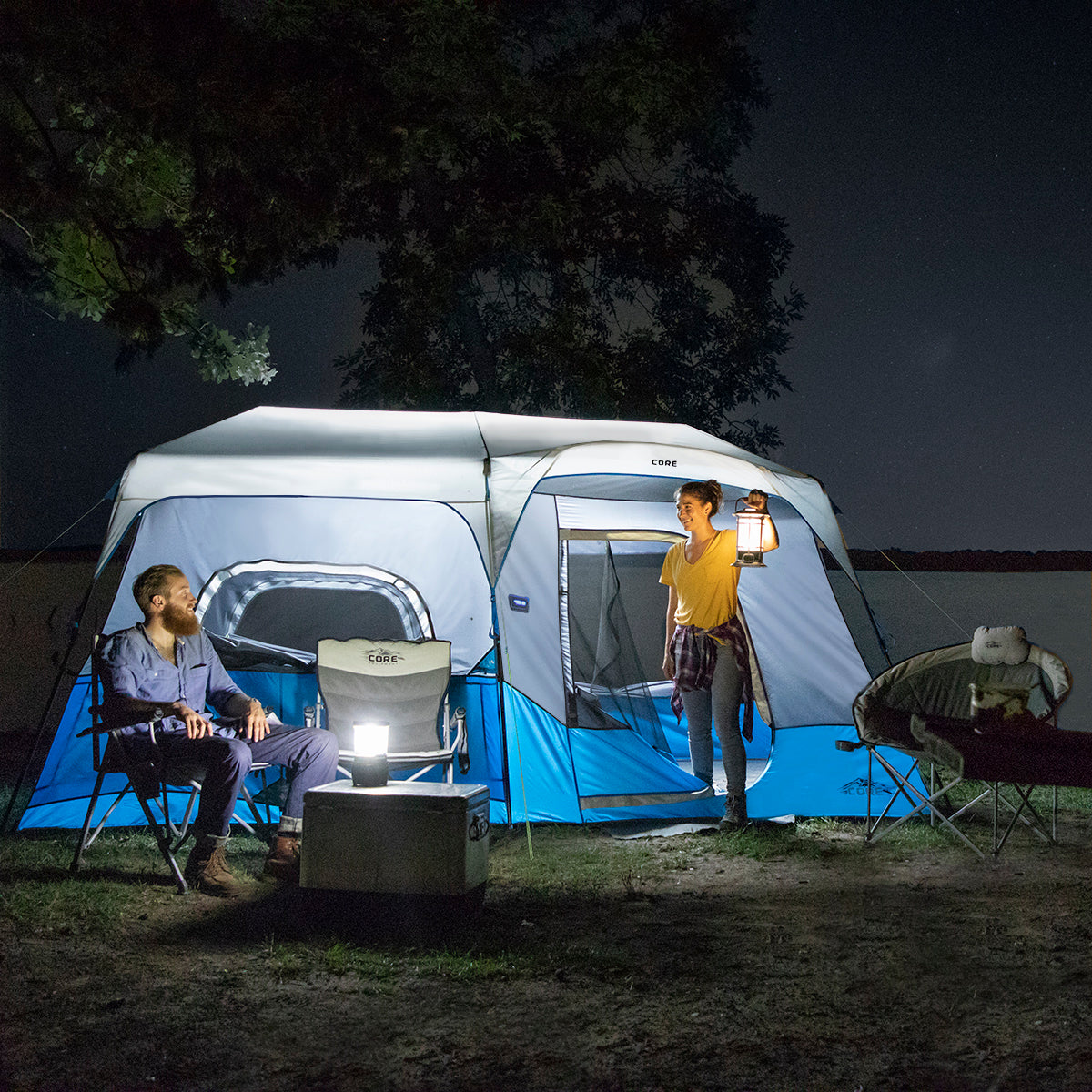 Two people are camping at night. One person is sitting on a chair outside a lit tent, holding a light. The other person is standing beside the tent, holding a lantern. Nearby, there is a cooler and another chair with a backpack on it. Trees and dark sky in the background.