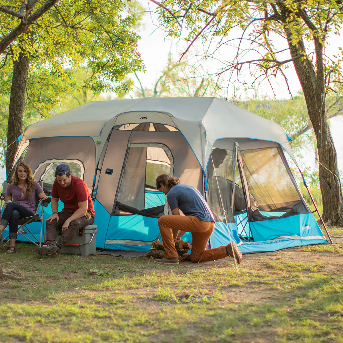 A group of four people is gathered outside a large blue and gray CORE 9 Person Lighted Instant Cabin Tent in a wooded campsite near a body of water. Two people are sitting on camping chairs while two others kneel and work on setting up the tent. The scene is bright and outdoorsy.