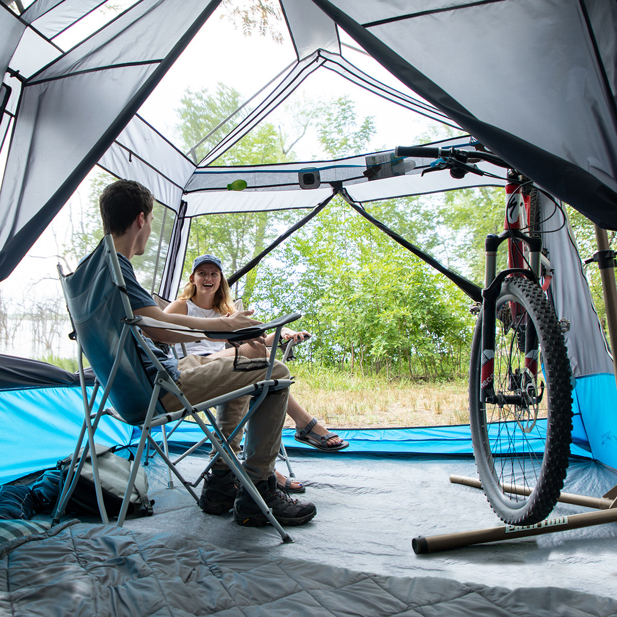 Two people are sitting in foldable chairs inside a spacious 9 Person Lighted Instant Cabin Tent. They are talking and smiling. A bicycle is positioned on the right side of the CORE tent. The tent door is open, revealing a lush, green outdoor setting with trees and grass.