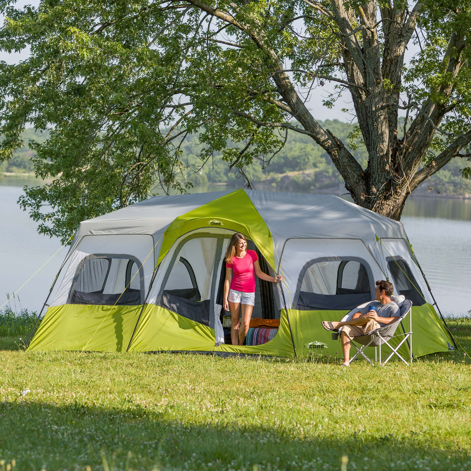 A woman stands at the entrance of a large, green and gray CORE 12 Person Instant Cabin Tent set up in a grassy area near a lake. A man sits in a folding chair nearby, looking at the woman. Trees and water are visible in the background under a clear sky.