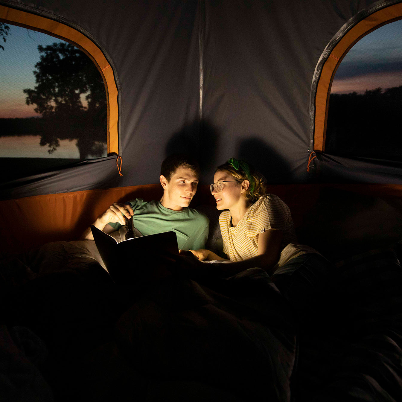 Two campers are using the flashlight to read a book inside a CORE tent.